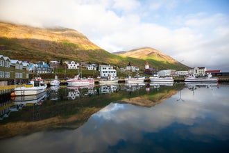 The hills, boats, buildings, and sky reflect onto the perfectly calm fjord waters of a village on the Trollaskagi (Troll Peninsula), part of Iceland's Arctic coastline.