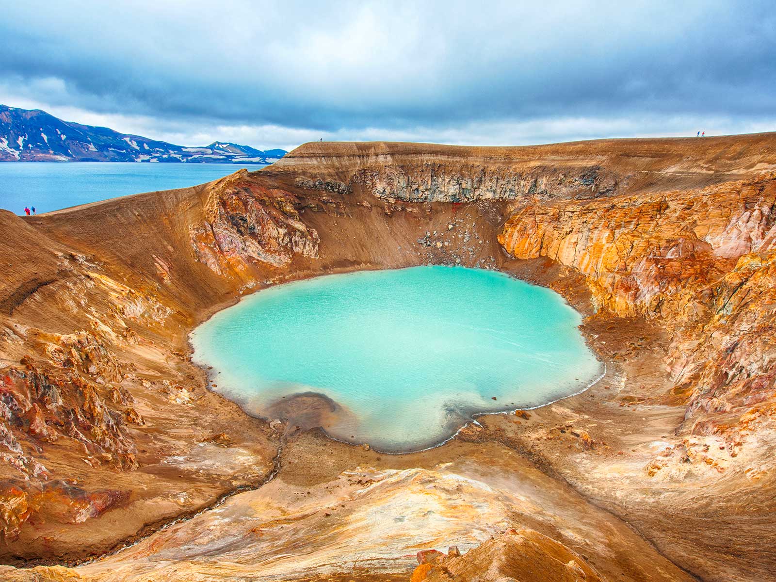 The milky-blue waters of the Viti crater lake are surrounded by brown volcanic rock with a glimpse of a large lake and mountains beyond.