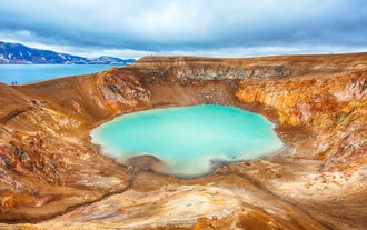 The milky-blue waters of the Viti crater lake are surrounded by brown volcanic rock with a glimpse of a large lake and mountains beyond.