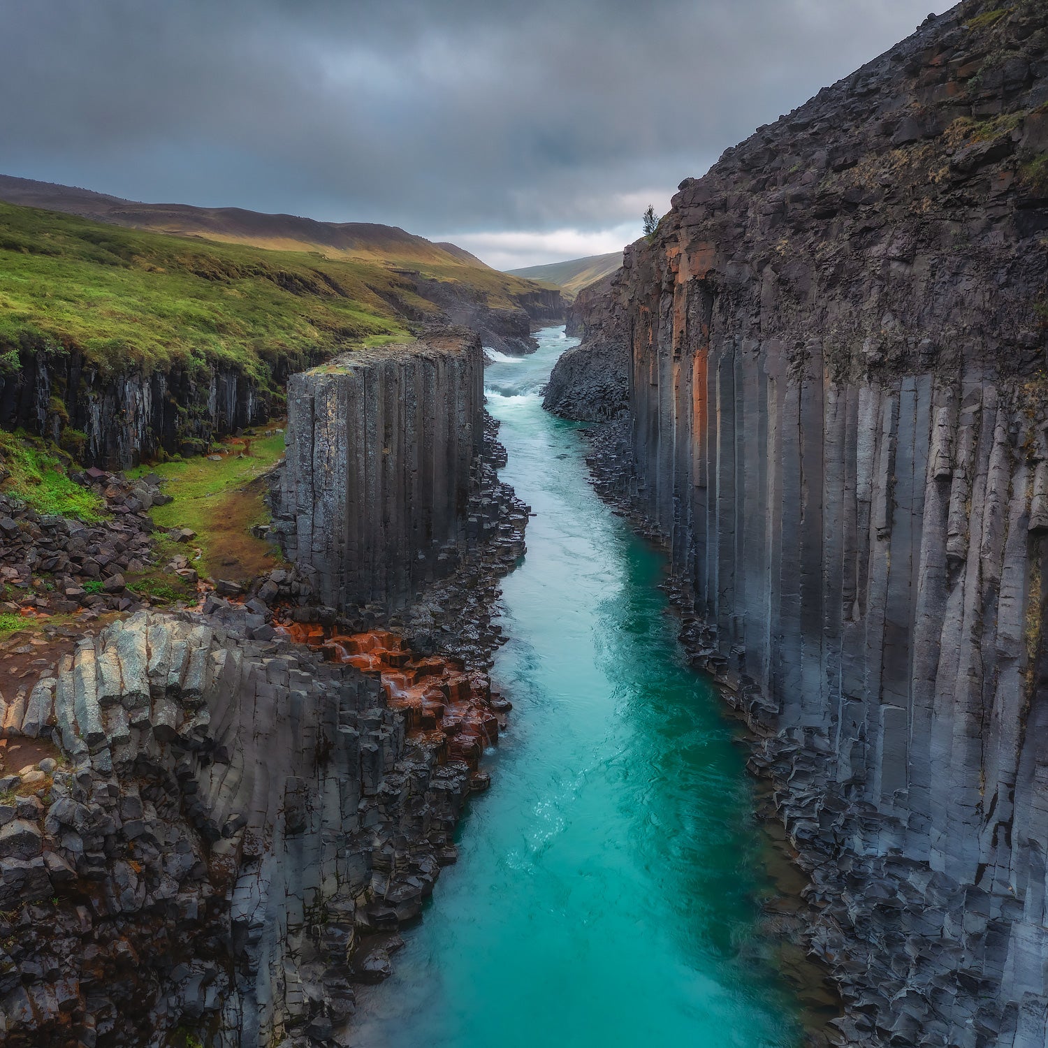 Basalt canyon of Studlagil in East Iceland with turquoise river flowing through dramatic columnar lava formations on both sides, captured under cloudy skies.