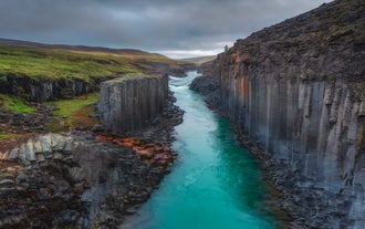 Basalt canyon of Studlagil in East Iceland with turquoise river flowing through dramatic columnar lava formations on both sides, captured under cloudy skies.