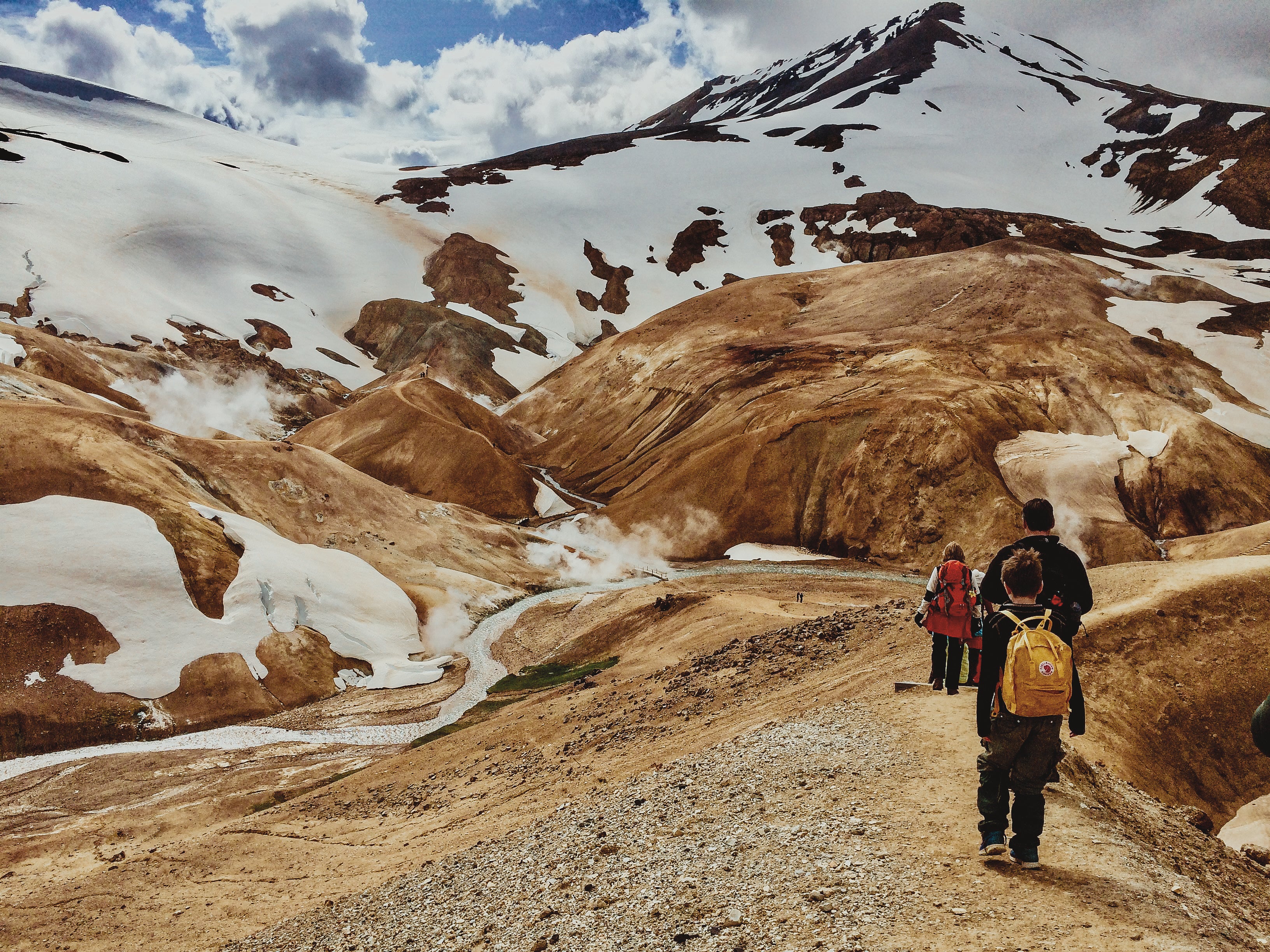 Hikers walking through Hveradalir geothermal valley in Kerlingarfjoll, with steaming vents, red hills, and patches of summer snow