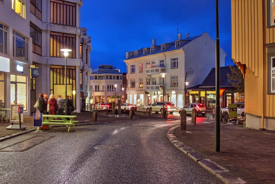 Evening view of a street in downtown Reykjavik, Iceland, with people walking, cars passing, and buildings lit under a deep blue sky.