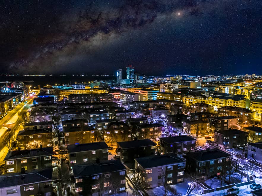 Nachtelijk luchtpanorama van de stadslichten en wijken van Reykjavik onder een sterrenhemel.
