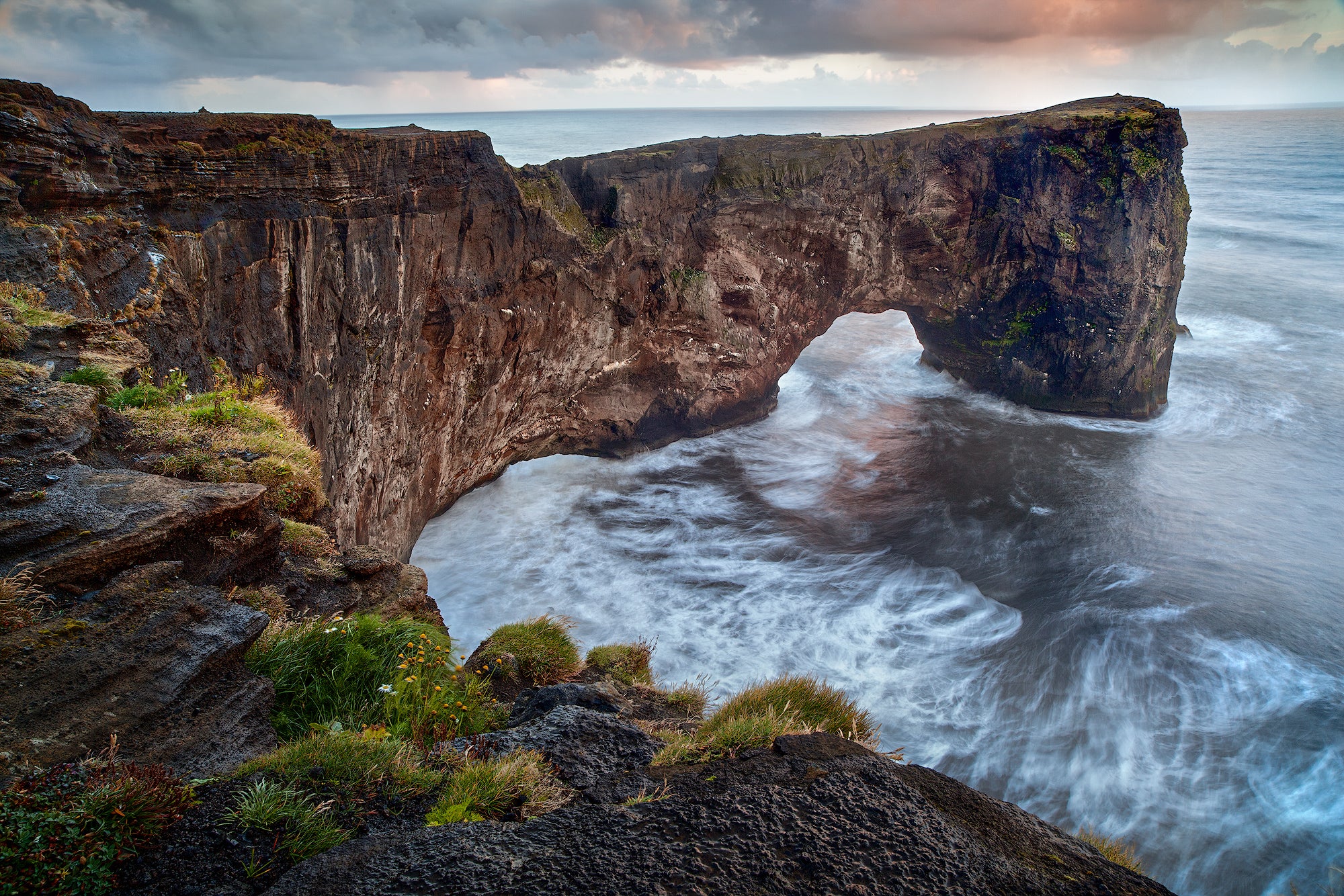 The Dyrholaey Promontory stretches into the ocean under a cloudy summer sky, with green cliffs and dramatic coastal views.
