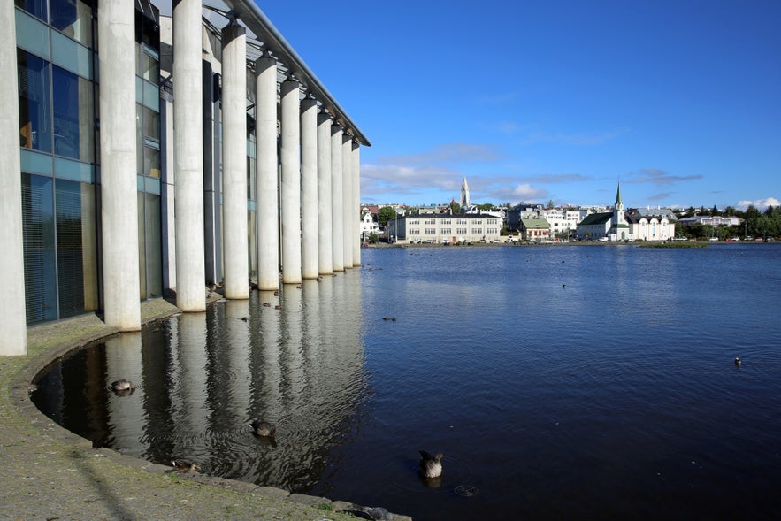 Reykjavik City Hall columns reflected in Tjornin Pond, with downtown Reykjavik skyline and birdlife in the background