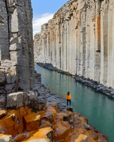 A traveler stands beside the blue glacial river and basalt columns of Studlagil Canyon in East Iceland.