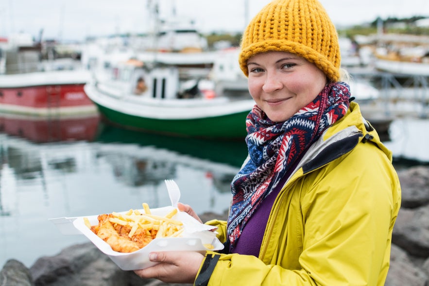 Young woman in yellow raincoat eats a portion of fish and chips in the port of Husavik, Iceland.