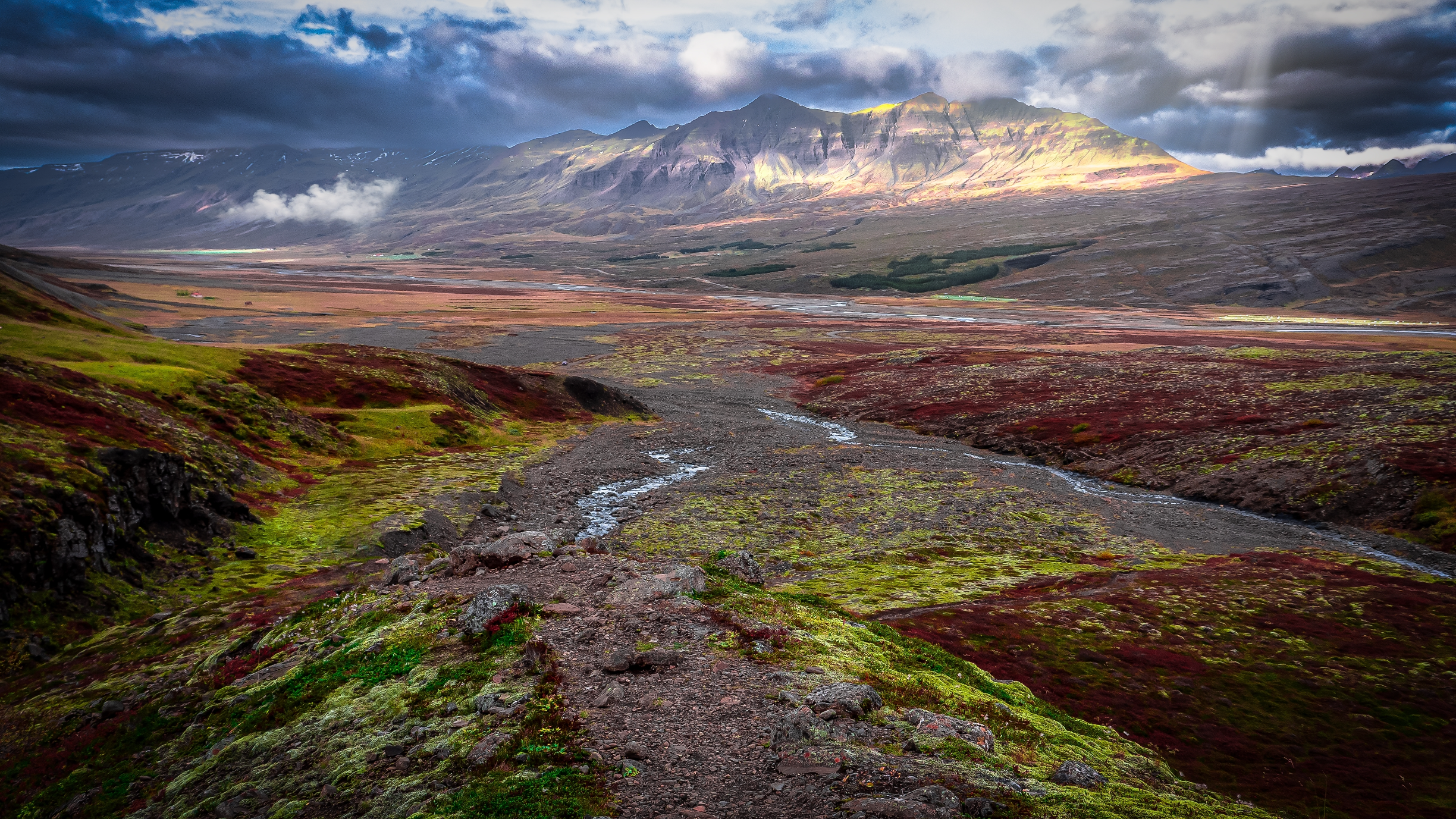 A stream winds through Flugufoss Valley in East Iceland, with sunlight illuminating the distant mountains.