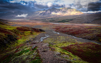 A stream winds through Flugufoss Valley in East Iceland, with sunlight illuminating the distant mountains.