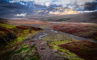 Un ruisseau serpente à travers la vallée de Flogufoss en Islande de l’Est, tandis que le soleil illumine les montagnes au loin.
