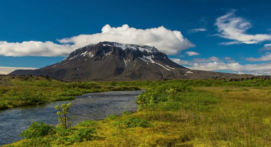 Snow-capped Mount Herdubreid rises above a lush green valley with a winding river under a clear blue sky in Iceland.