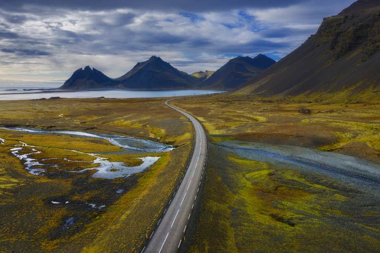A scenic aerial view of Route 1 in Iceland, showcasing a long road stretching through a diverse landscape of mountains, rivers, and coastal areas in the Eastfjords.