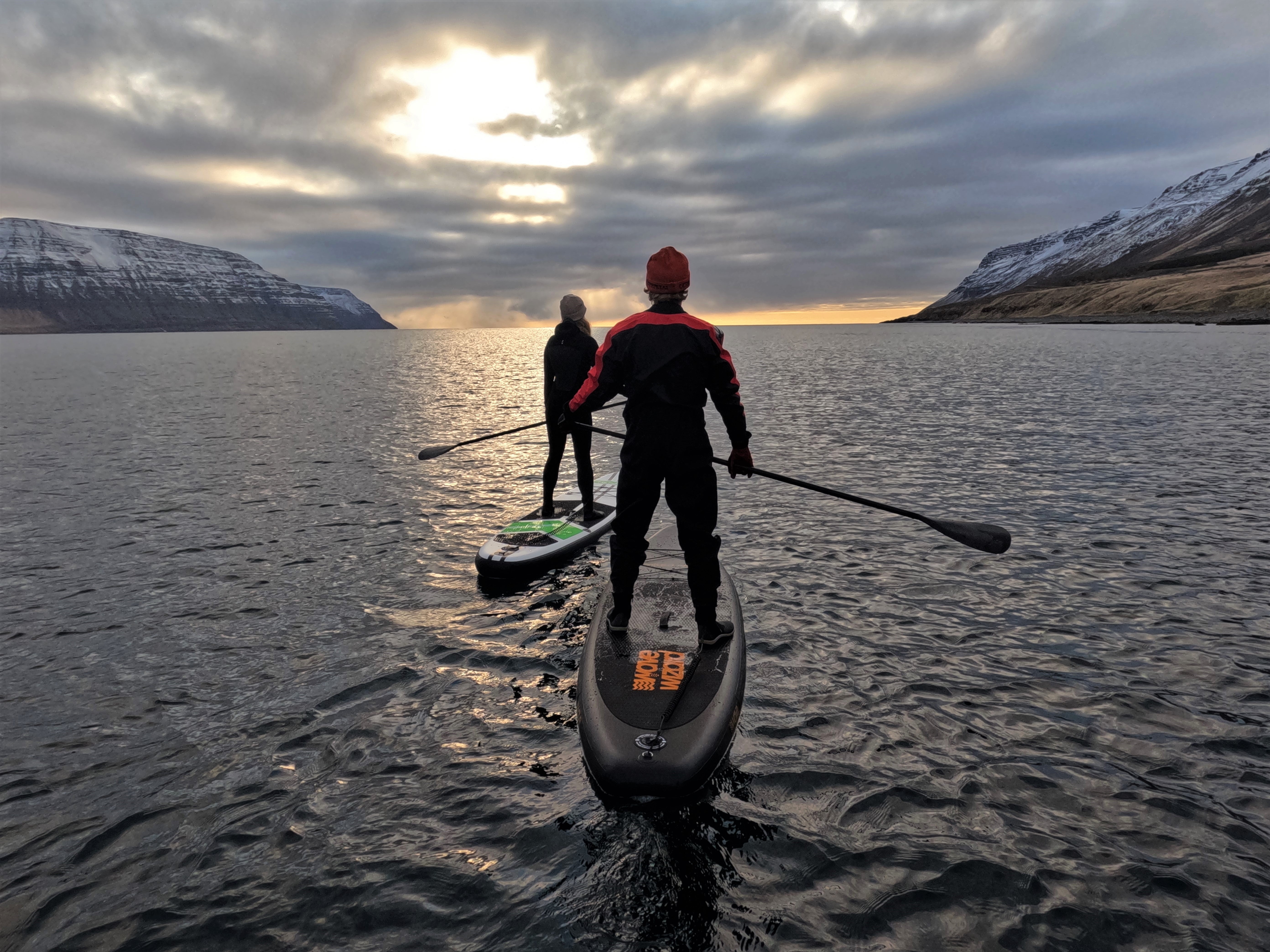 Two people enjoying Iceland paddleboarding on a calm fjord at sunset with mountains in the background.