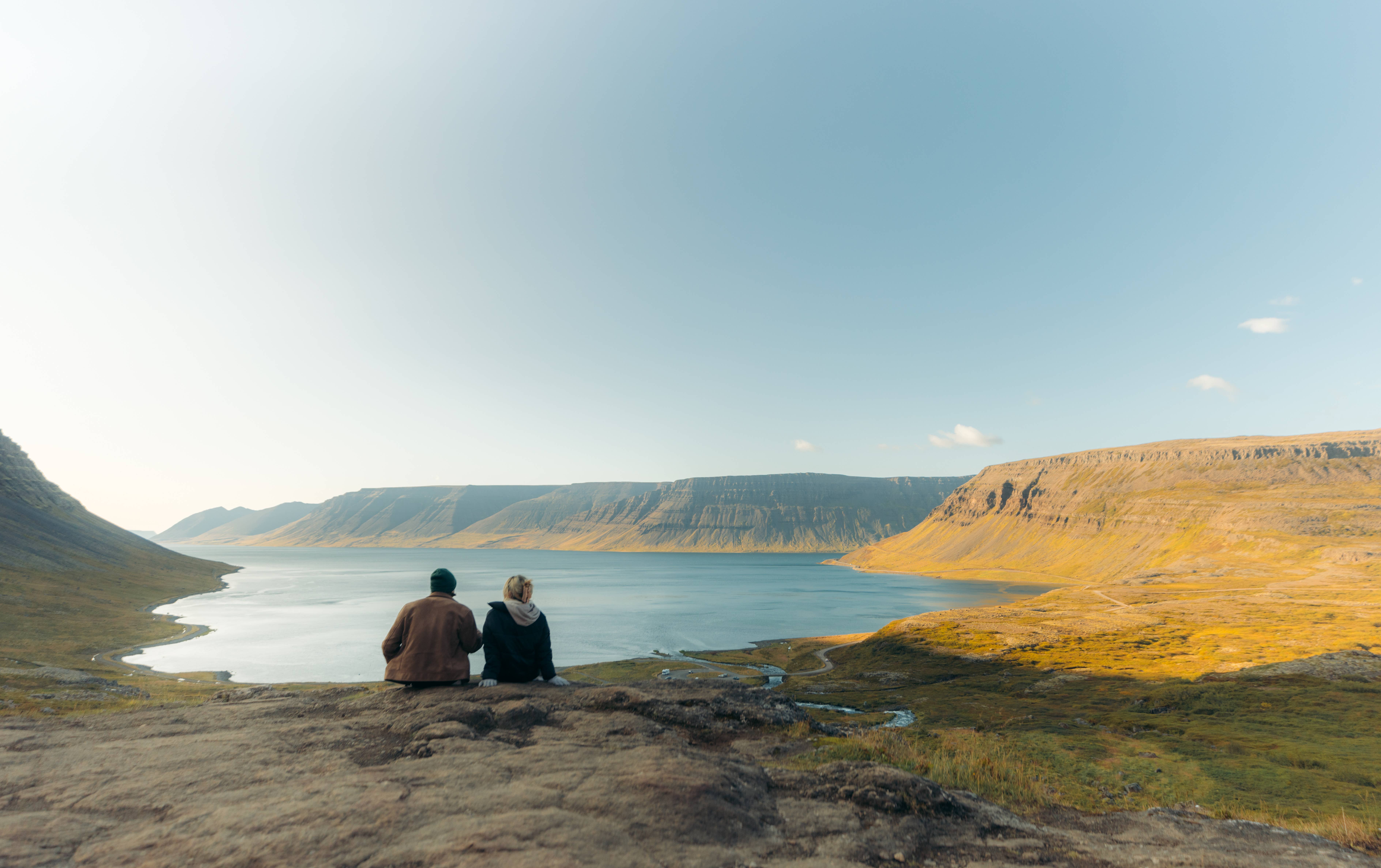A man and a woman sit on the edge of the fjord, looking out over the water toward distant mountains during a Westfjords tour.