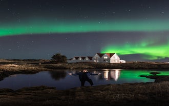A man stands under the northern lights with a frozen lake and white houses in the background.