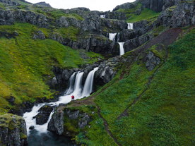 Klifbrekkufossar Waterfall in Iceland, showing multiple cascades of water flowing down a lush green, rocky mountainside with two people standing near the base.