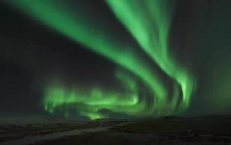 Green northern lights swirl brightly across a dark Icelandic sky above snowy fields.