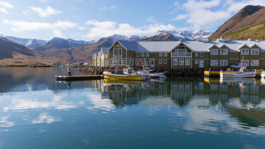 Scenic view of the harbor in Siglufjordur, Iceland, with traditional green wooden buildings and fishing boats docked on calm, reflective water. Snow-capped mountains rise in the background under a blue sky with scattered clouds.