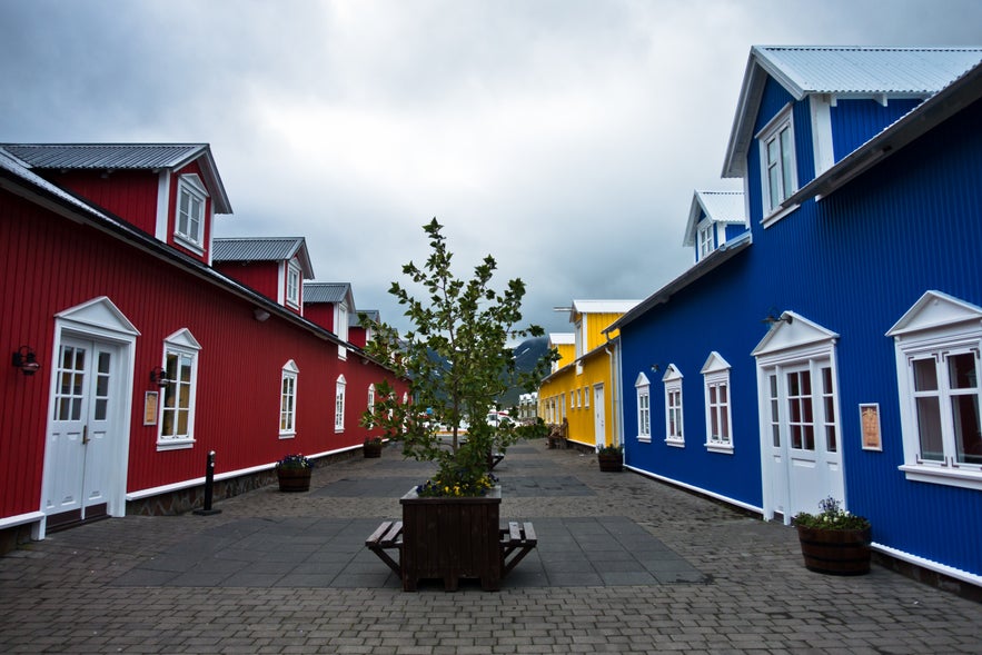 A colorful side street just off Adalgata in Siglufjordur, Iceland, featuring vibrant wooden buildings painted red, blue, and yellow. The charming facades have white-trimmed windows and doors.