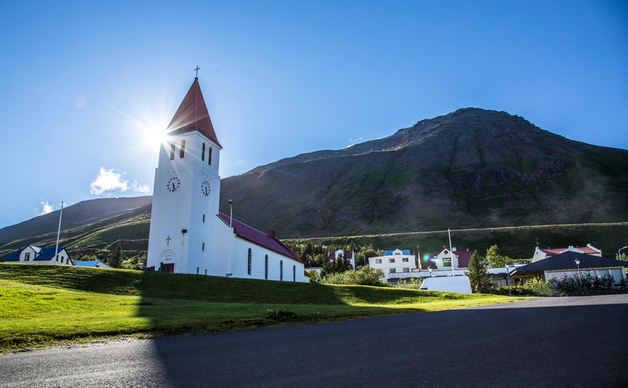 Siglufjordur Church in Iceland, a white wooden church with a red roof and steeple, stands on a grassy hill with the sun shining brightly behind its clock tower.