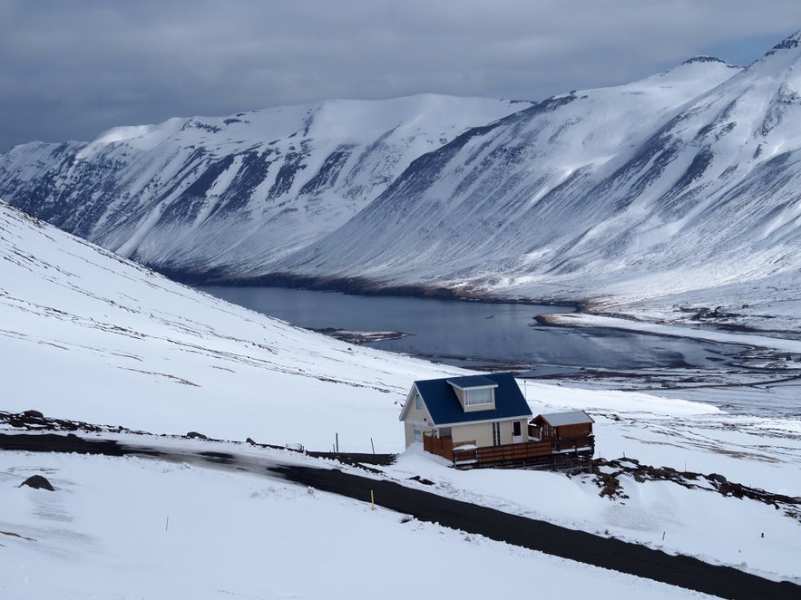 Winter view over Siglufjordur fjord in North Iceland, with snow-covered mountains and a calm, partially frozen body of water below. A small house with a blue roof sits on a slope in the foreground.
