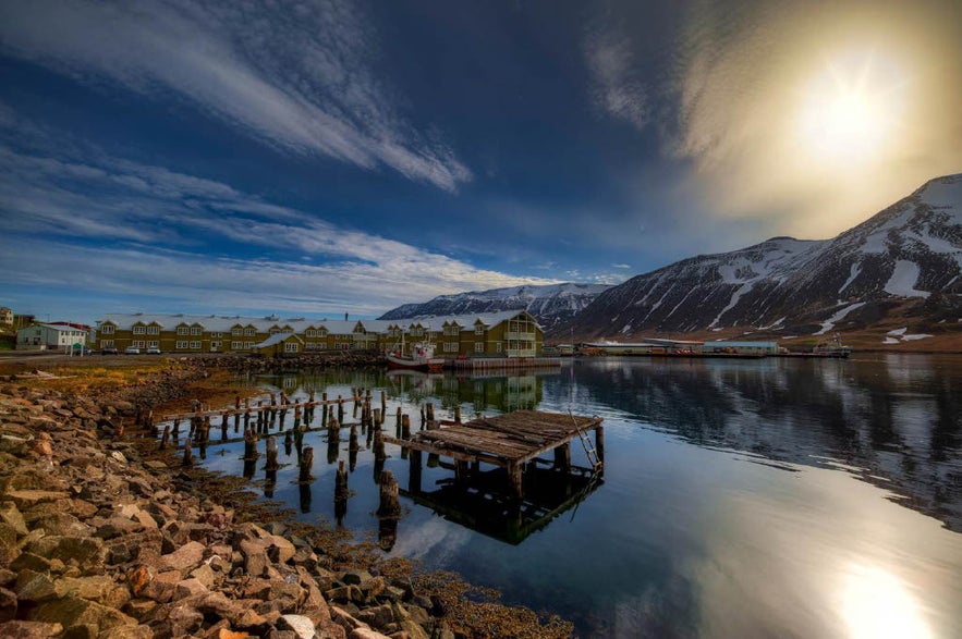 Harbor view of Siglufjordur under the warm glow of the low Arctic sun, nearing the midnight sun season. The calm water reflects green waterfront buildings, moored fishing boats, and the remains of an old wooden pier, against snow-striped mountains and a richly colored sky.