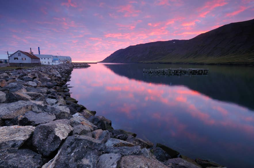 Vibrant twilight over Siglufjordur fjord in Iceland, with pink and purple clouds reflecting on the still water. A rocky shoreline leads to old industrial buildings and a decaying wooden pier.