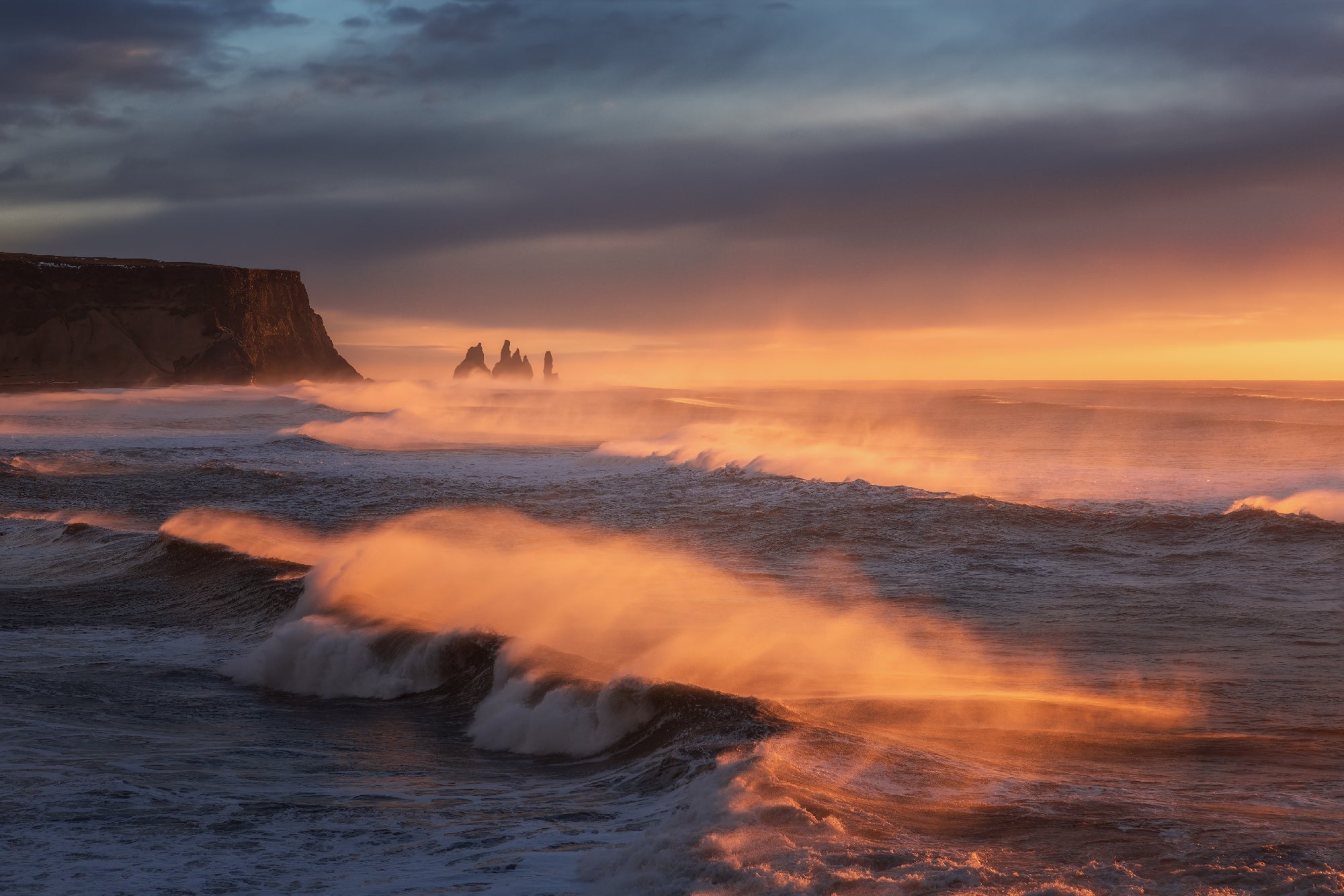 Powerful waves of the Atlantic crash against Reynisfjara beach with a sea stack slightly obscured from view.