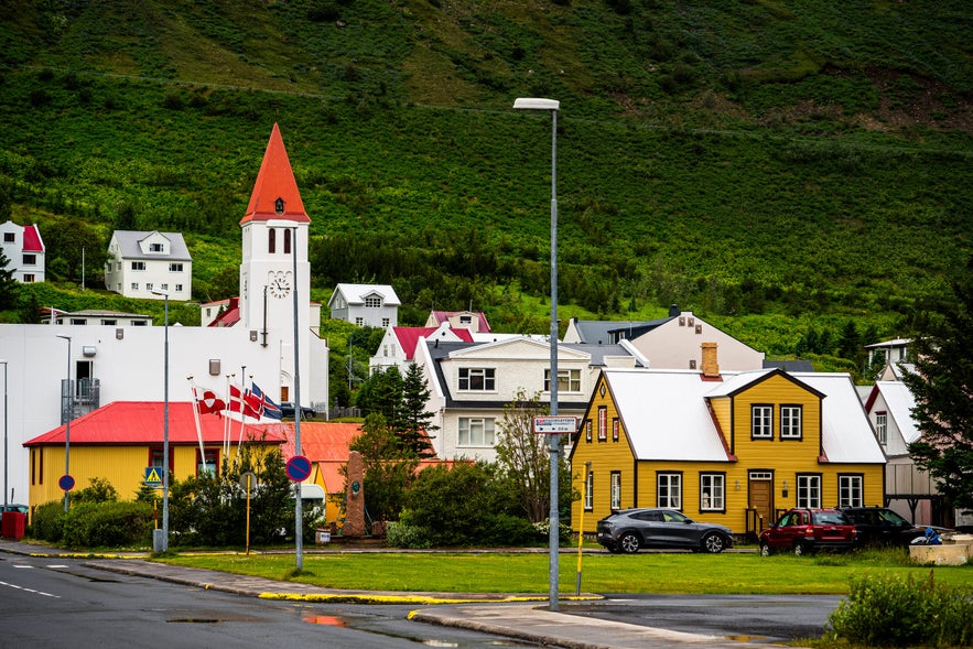 Colorful buildings in central Siglufjordur, Iceland, including the Folk Music Centre. In the background, the white church with a red steeple stands out against the lush green hillside, while Nordic flags wave beside a red-roofed building.