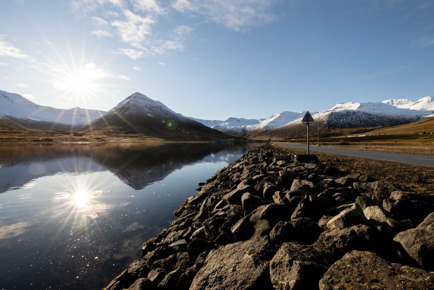 Scenic view of a mountain road near Siglufjordur, Iceland, with a calm lake on one side and snow-capped peaks in the distance. The sun casts a bright glare and long shadows, while a rocky embankment runs alongside the asphalt road.