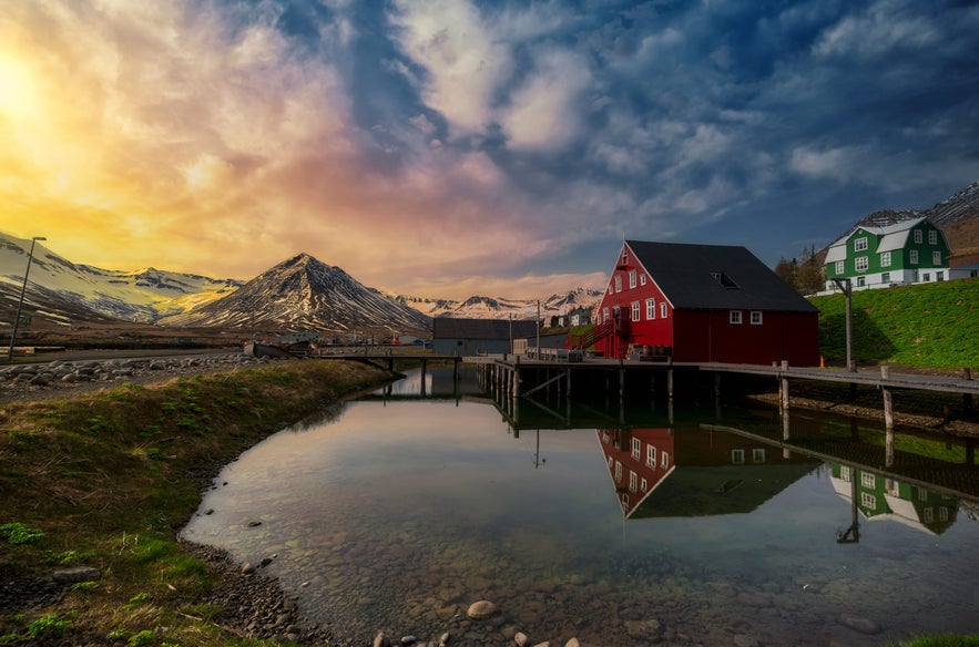 Stunning view of Siglufjordur, Iceland, with the iconic red Roaldsbrakki building from the Herring Era Museum reflected in calm water. Snow-capped mountains rise in the background under a vibrant sky transitioning from golden sunset tones to deep blue.