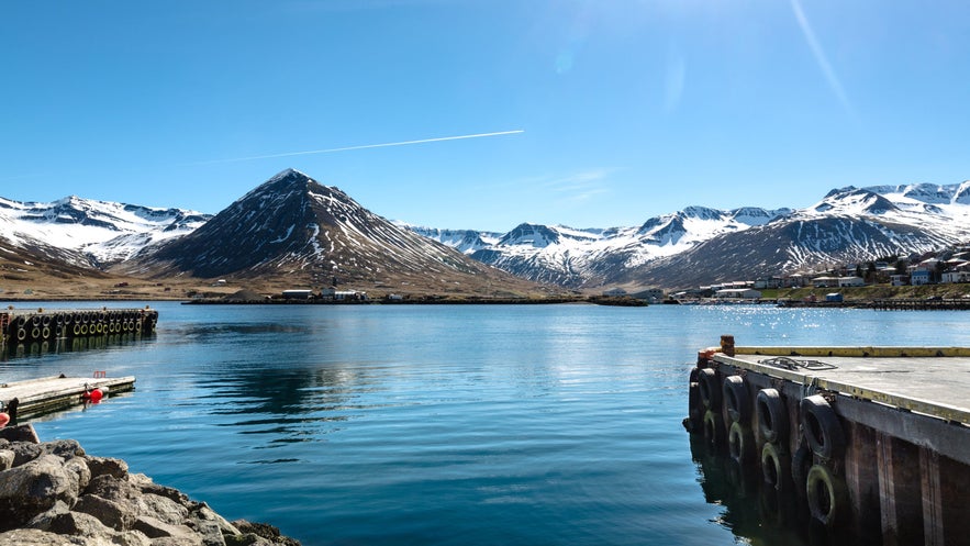 Clear, sunny view of Siglufjordur fjord in North Iceland, with sparkling blue water surrounded by snow-capped mountains. A distinctive pyramid-shaped peak rises at the center, while docks with tires line the harbor edge in the foreground. 