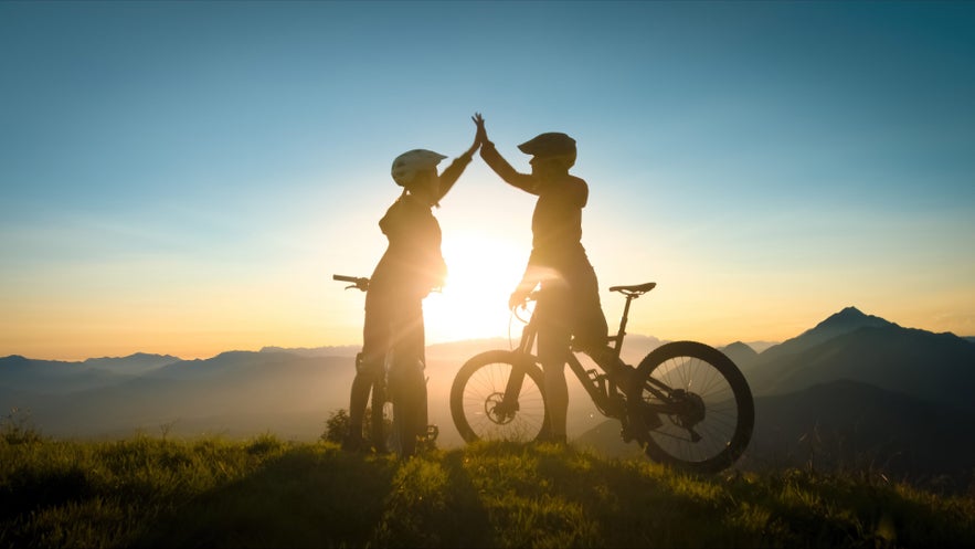 Silhouettes de deux cyclistes se faisant un high five au coucher du soleil sur une crête près de Siglufjordur, Islande. Leurs vélos reposent à côté alors que le soleil projette une lumière dorée sur la colline.