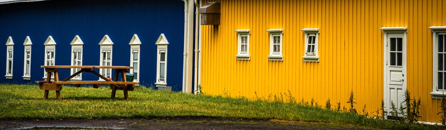 Close-up view of the blue and yellow buildings at the campsite in the town center by the harbor in Siglufjordur, Iceland. The structures feature white-trimmed windows and corrugated metal siding, with a wooden picnic bench on the grassy area between them.