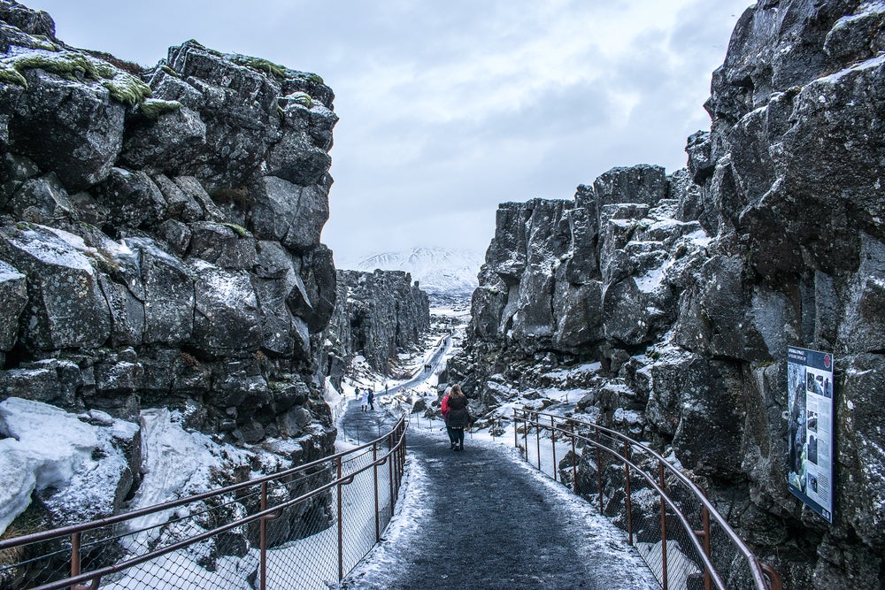 Visitors walk along a path through the snowy, dramatic rock formations of Thingvellir National Park in Iceland during winter.