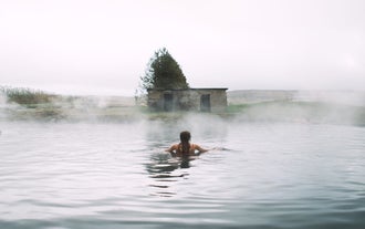 A woman relaxes in the misty, steaming waters of the Secret Lagoon in Iceland, with a small building and a tree visible on the shore.