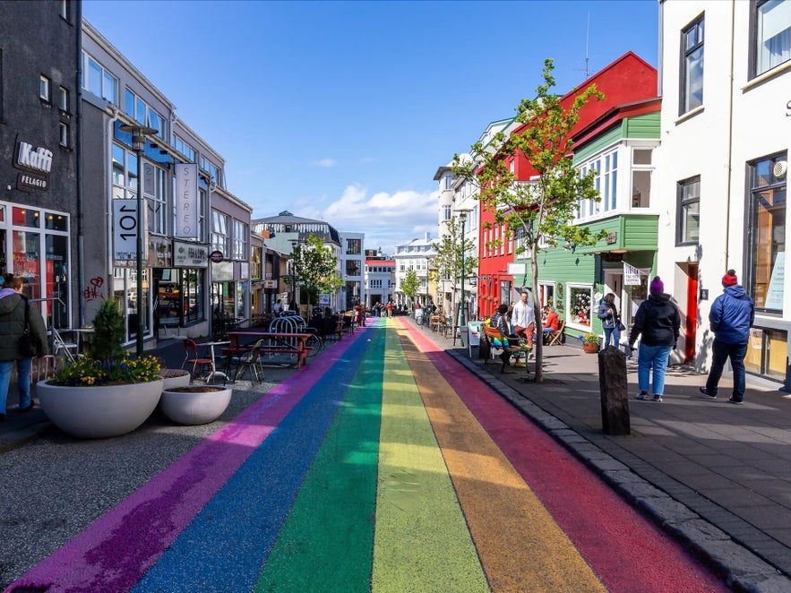 Colorful rainbow-painted Rainbow Street in downtown Reykjavik, Iceland, with vibrant buildings, cafes, and pedestrians enjoying a sunny day.