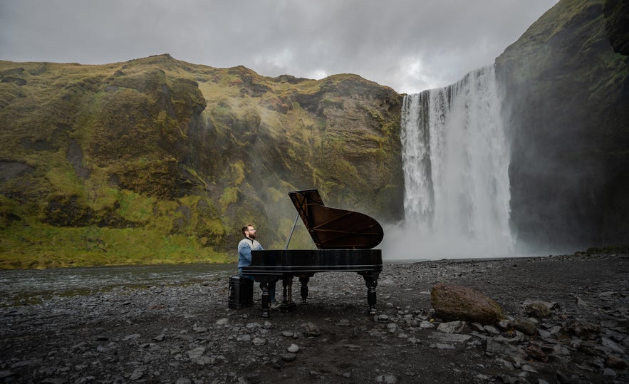 Pianist performing on a grand piano in front of Sk&oacute;gafoss waterfall in Iceland.