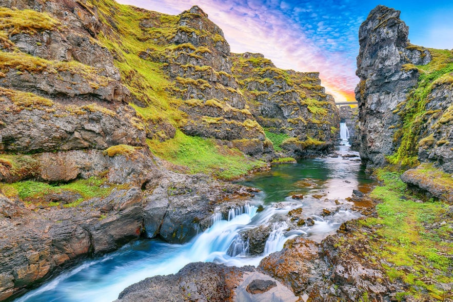 Kolagljufur canyon in Iceland, with moss-covered cliffs, a calm river, and a small waterfall beneath a bridge at sunset.