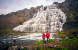 Two people in red jackets are standing near the base of Dynjandi Waterfall, surrounded by flowing water and rocky terrain.