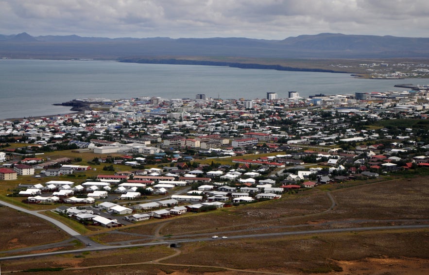 Aerial view of Keflavik, showing coastal town layout with residential neighborhoods, marina, and distant mountains under cloudy skies.