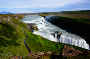 Gullfoss Waterfall cascading into a canyon with a rainbow forming over the mist and visitors walking along the path.
