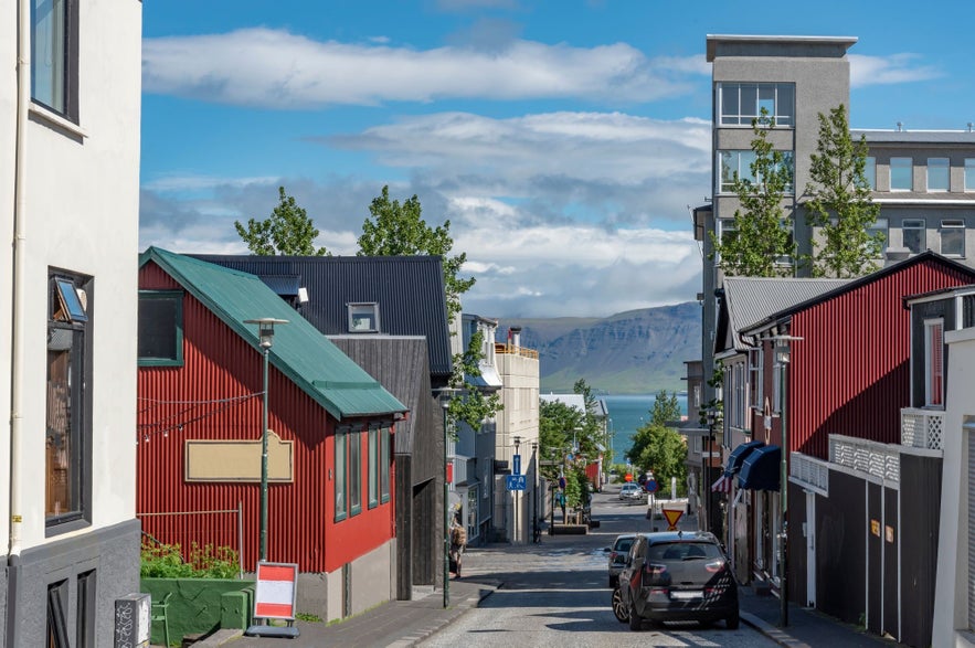 Street view in downtown Reykjavik, Iceland, featuring colorful houses, parked cars, and a distant view of Mount Esja and the ocean on a clear summer day.