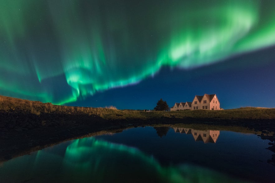 Aurora borealis above rural houses with water reflections, highlighting the unique charm of real estate in Iceland.
