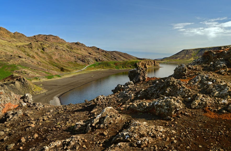 Volcanic landscape with rugged lava rocks, a winding shoreline, and still water at Kleifarvatn Lake on the Reykjanes Peninsula in Iceland, under clear blue skies.