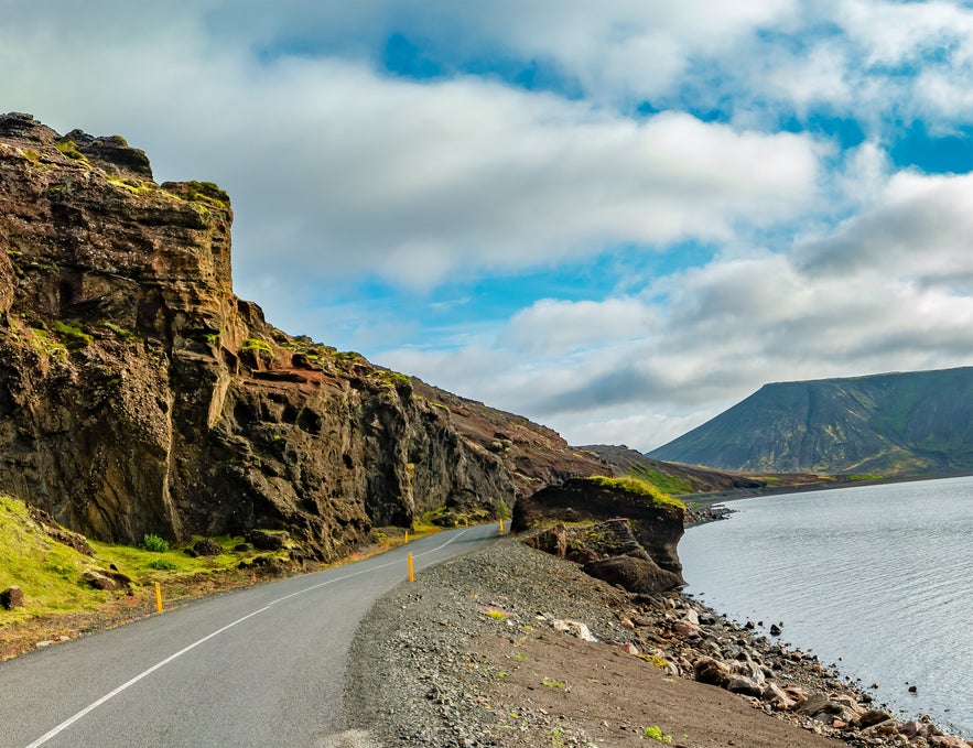 Scenic coastal road winding along the cliffs of Kleifarvatn Lake on Iceland’s Reykjanes Peninsula, with dramatic rock formations and a mountain in the background under partly cloudy skies.