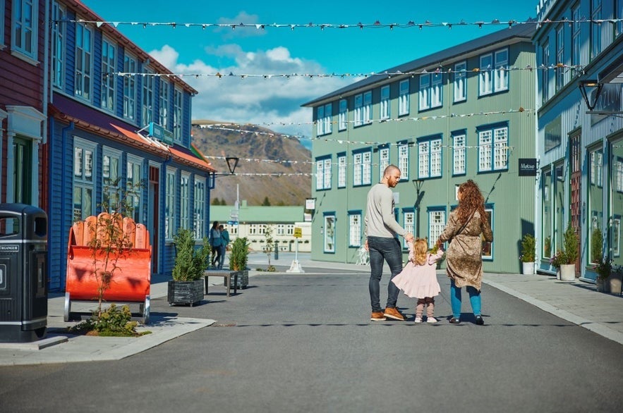 Family walking through the colorful New Old Town district in Selfoss, Iceland, with renovated buildings, string lights, and mountain views under a clear blue sky.