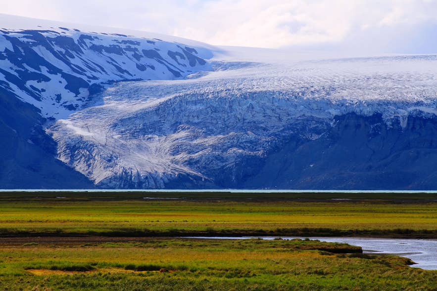 Uitzicht op de dramatische ijsrand en ruige kliffen van de Langjökull-gletsjer in IJsland, met groene graslanden op de voorgrond en een heldere, deels bewolkte lucht.