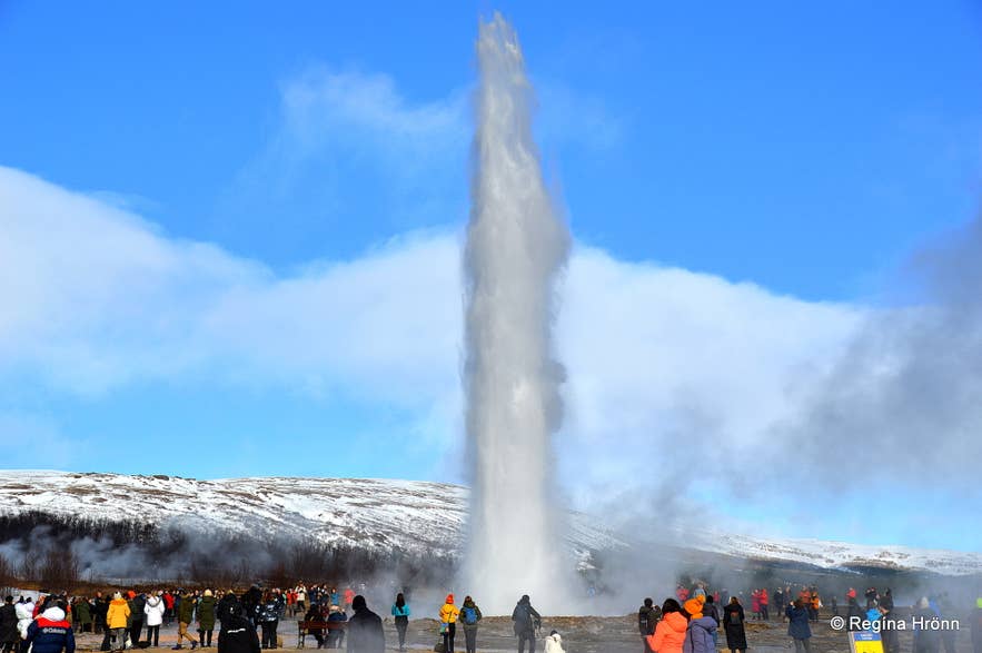 Crowd watching Strokkur erupt against snowy hills on a clear March day in the Geysir area.